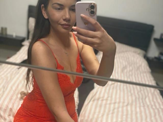 MoonlightCat, sensuelle dans une robe rouge en dentelle, assise sur un lit, prenant un selfie avec son téléphone, montrant ses seins et un léger sourire, avec de longs cheveux bruns..