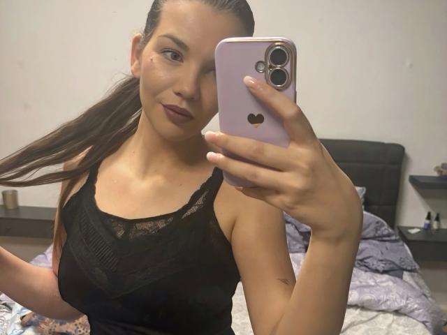 MoonlightCat, une jeune femme aux longs cheveux bruns, portant un haut en dentelle noire, prend un selfie devant un miroir avec un téléphone rose, montrant son petit tatouage sur son bras gauche, et porte un rouge à lèvres foncé..
