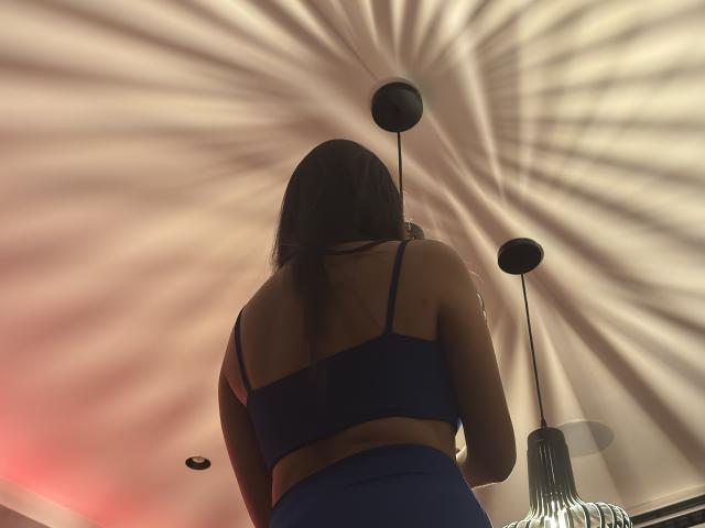 NyraMoon, con lunghi capelli castani, indossa un reggiseno blu e un paio di pantaloni a vita alta dello stesso colore, ed è in piedi con la schiena rivolta verso la fotocamera, sotto un soffitto con motivi luminosi radiali..