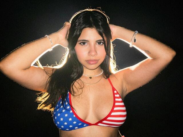 CelesteMorel with hands in dark hair, wearing an American flag bikini, highlighting her tits and small ass, against a black background, with a silver bracelet and necklace.