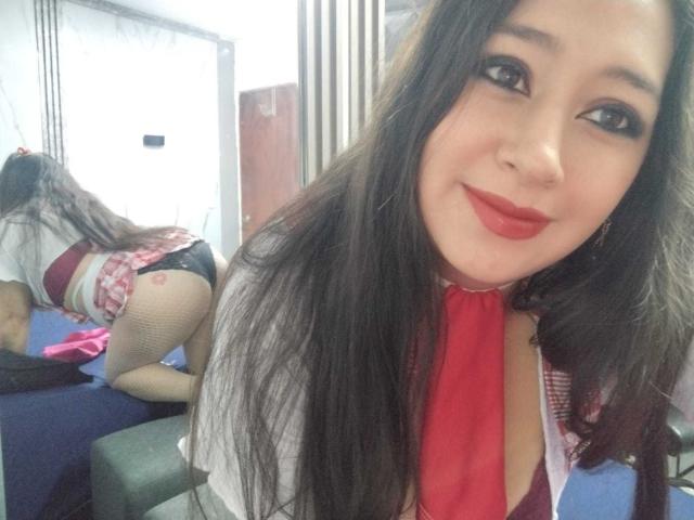 MelodyRoss, com longos cabelos pretos, batom vermelho e meias de rede, vestindo uma gravata vermelha e uma saia xadrez rosa, exibindo seu traseiro em uma selfie no espelho..