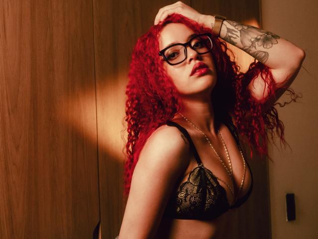 LilJane, com cabelo ruivo encaracolado, sutiã de renda preto, óculos e tatuagens, posando com a mão na cabeça, mostrando os seios e batom vermelho, usando um colar de ouro..