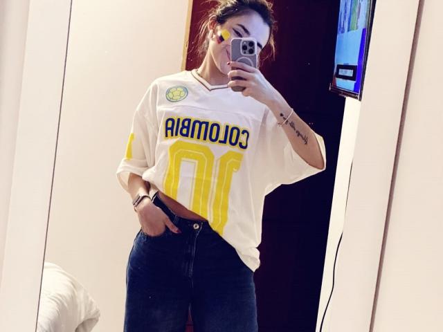 AdharaCooperr in een wit AlamoJoe shirt, met gele cijfers, en blauwe jeans, hand in de zak, spiegelzelfportret, telefoon in de hand, tatoeage op de pols, witte muur op de achtergrond..