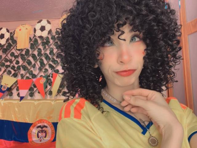 LaurenEvie69 with curly black hair, green eyes, red lipstick, nose ring, wearing yellow soccer jersey with orange stripes, hand near face, necklace, in colorful room.