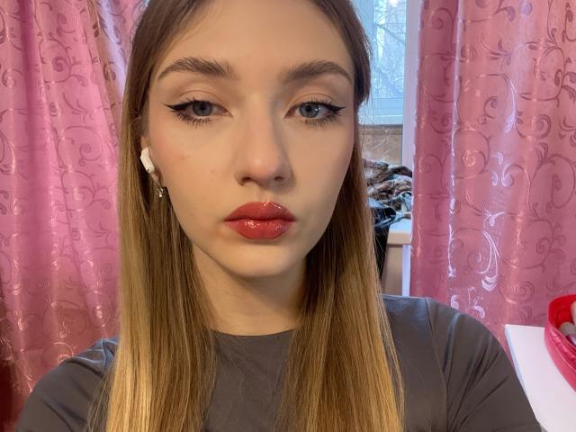 DenisseReins hat glänzende, rote Lippen, schwarzen Eyeliner mit Flügeln und langes, blondes Haar. Sie trägt ein graues Oberteil, im Hintergrund sind rosa Vorhänge und ein weißes Fenster zu sehen..