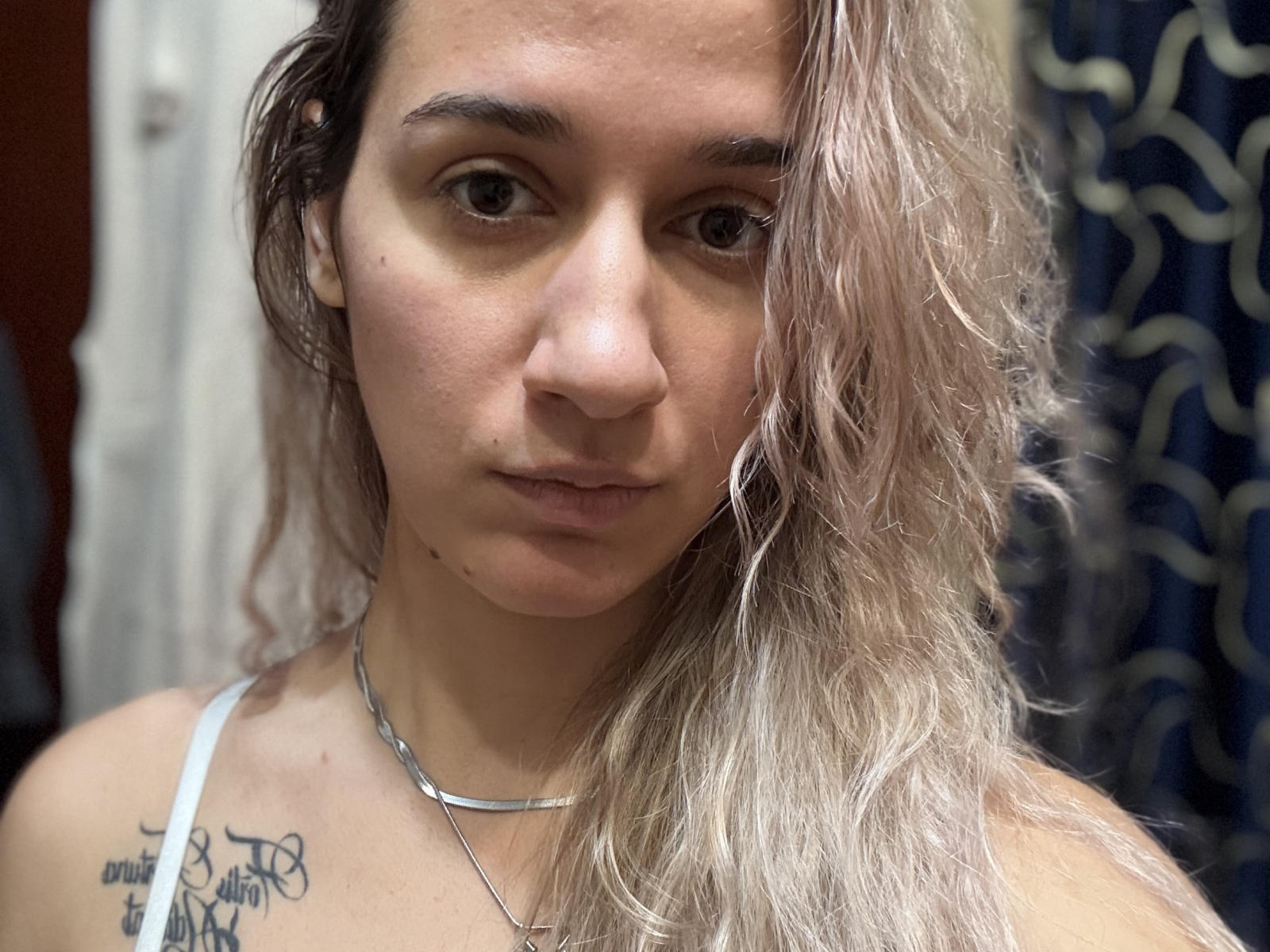 EthelBiangone mit welligem, blond-braunem Haar, heller Haut, kleiner Nase, braunen Augen, einem weißen Tanktop, einer Halskette und einem Tattoo auf der rechten Schulter, die direkt in die Kamera schaut..