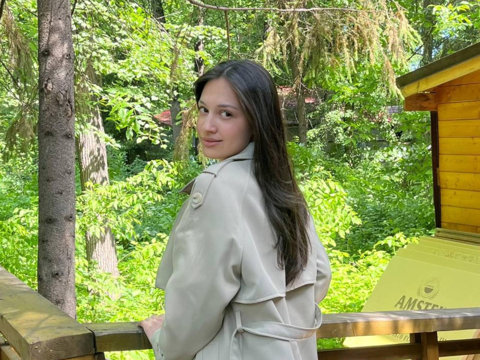 Adrianne Rossey, avec de longs cheveux foncés, portant un manteau beige, se tient sur un balcon en bois, regardant en arrière avec un léger sourire. On aperçoit une forêt verdoyante et une maison en bois jaune en arrière-plan..