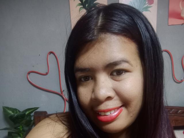 MariahSquirting69 ha i capelli neri e lisci, rossetto rosso e un leggero sorriso, con un filo rosso a forma di cuore sullo sfondo e una pianta verde parzialmente visibile..