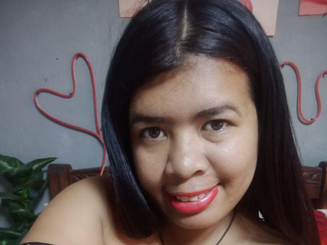 MariahSquirting69, con i capelli neri, il rossetto rosso e un sorriso carino, guarda la fotocamera. Ha un seno di medie dimensioni e un viso leggermente lentigginoso. Decorazioni a forma di cuore rosse appese al muro..