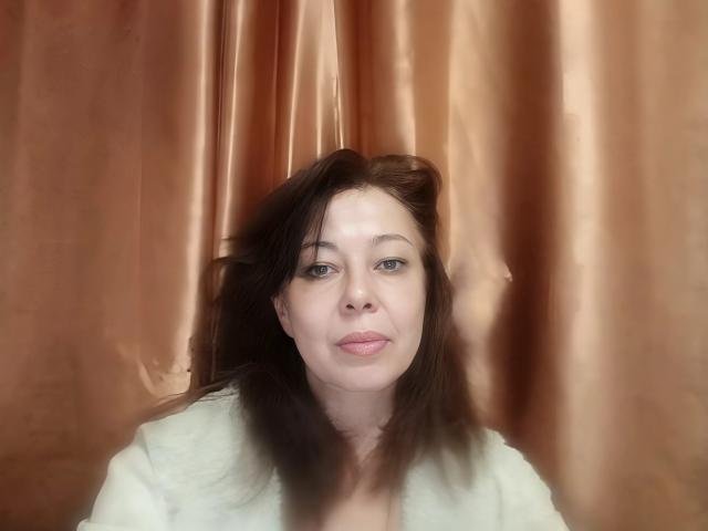 Priya Mayes, con su cabello castaño peinado hacia un lado, piel pálida, ojos azules y labios rosados, vestida con una blusa blanca, con un fondo de cortina marrón borroso..