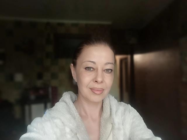Priya Mayes, con un sorriso adorabile, indossa un morbido accappatoio bianco, ha la pelle chiara, gli occhi marroni e i capelli castani raccolti. Ha il seno piccolo e sta guardando la fotocamera..
