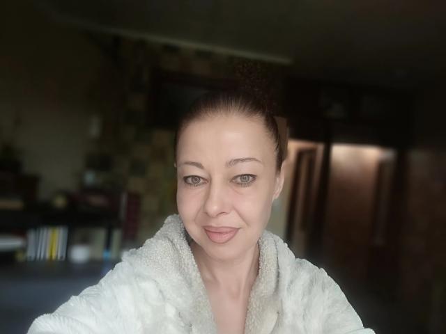 Priya Mayes, con la pelle chiara, gli occhi marroni e i capelli castani raccolti in uno chignon, indossa un morbido accappatoio bianco e sorride dolcemente alla fotocamera. Si può notare il suo piccolo naso, leggermente rivolto verso l'alto..