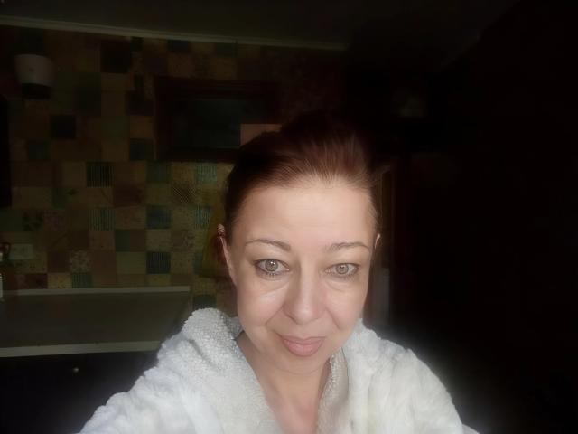 Priya Mayes, avec une peau claire, des cheveux bruns et des yeux verts, souriant légèrement, portant une serviette blanche et moelleuse, dans une salle de bain peu éclairée..