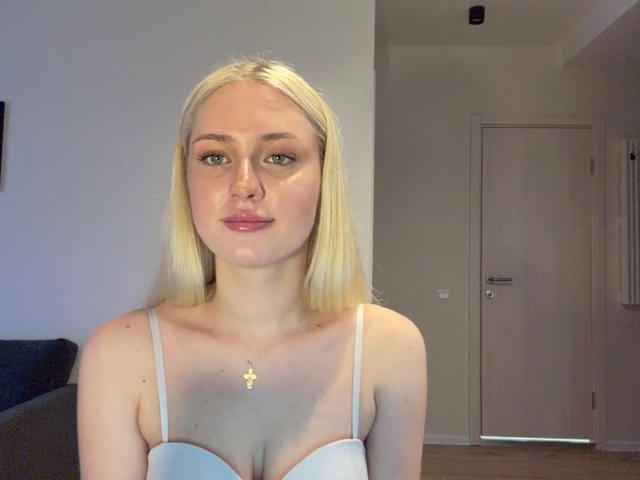 NancyBijou, com cabelo loiro, lábios carnudos, vestindo um vestido branco com alças, mostrando seios pequenos, pele clara, olhos verdes e um colar de ouro, em um quarto simples..