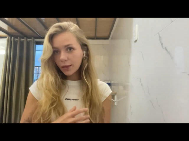 Die blonde LennaTjandra mit welligem Haar, die ein weißes T-Shirt trägt, hat ein süßes Gesicht, kleine Brüste und berührt mit einer Hand ihre Brust, während sie in die Kamera schaut..