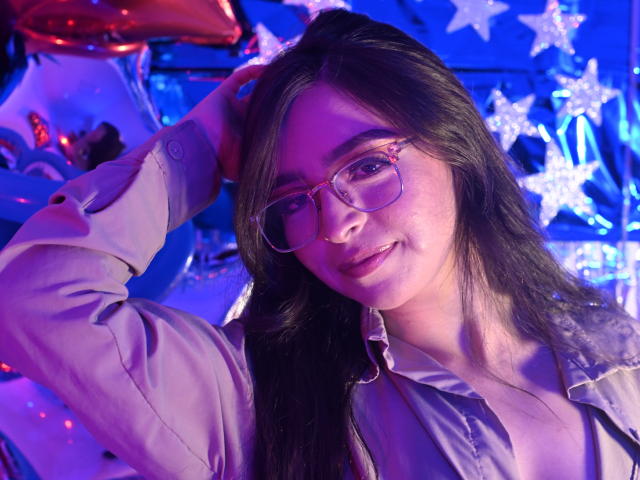 EvaMcAllister with glasses, long dark hair, pink shirt, hand in hair, smiling, blue and purple lighting, looking at camera, slight blush, cute, attractive.