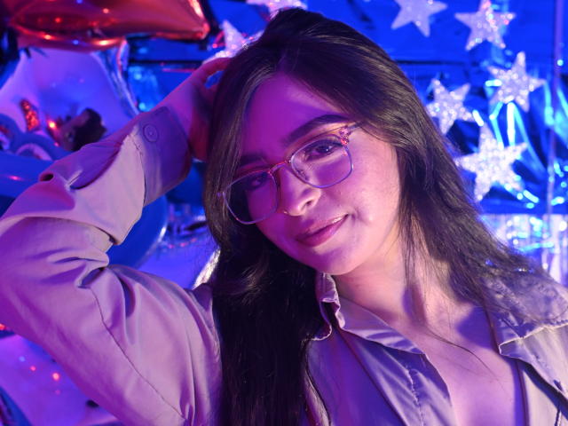 EvaMcAllister with long dark hair, wearing glasses and a beige shirt, raising her arm, showing her cute face and small tits, under blue and purple lighting.
