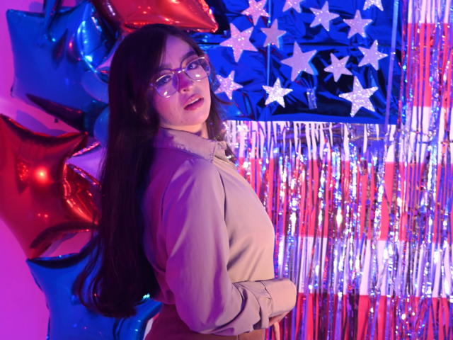 EvaMcAllister, met lang donker haar, een bril en een beige jas, staat met haar rug lichtjes gedraaid, kijkend over haar schouder. Rode en blauwe ballonnen, ster-vormige decoraties en zilveren slingers..