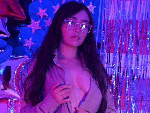 EvaMcAllister met lang donker haar, een bril en een roze shirt dat open staat om kleine borsten te laten zien, staat in paars en blauw neonlicht en houdt een shirt vast. Op de achtergrond zijn sterren en strepen te zien..