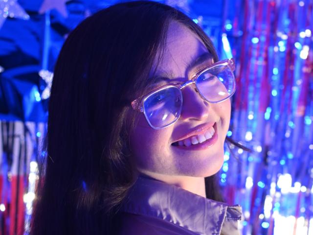 EvaMcAllister with long black hair, smiling, wearing clear plastic glasses, and a shiny gray jacket, blue and purple lights reflecting on her face.