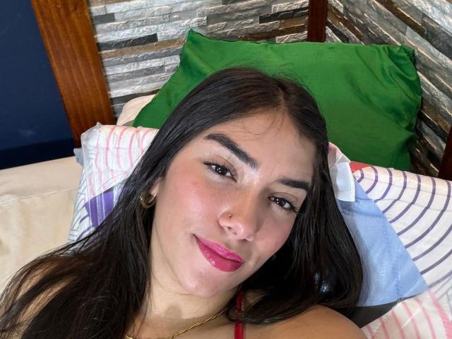 SharonKlart tem cabelos pretos longos, olhos castanhos, batom rosa e um leve sorriso. Ela está deitada em uma cama com travesseiros verdes e listrados, vestindo uma blusa vermelha..