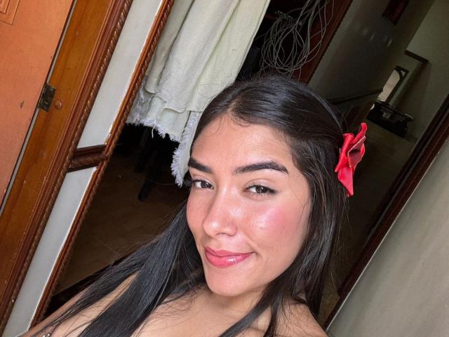 SharonKlart, com lábios rosa brilhantes, cabelo preto e liso, e um prendedor de cabelo vermelho, sorrindo para a câmera, exibindo seu rosto adorável e levemente corado..
