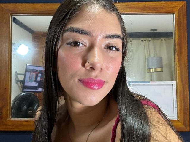 SharonKlart, con cabello negro largo, lápiz labial rosa y ojos marrones, vestida con una blusa roja, sonriendo ligeramente, reflejo en un espejo de madera, tono de piel medio, maquillaje sutil, pestañas negras..