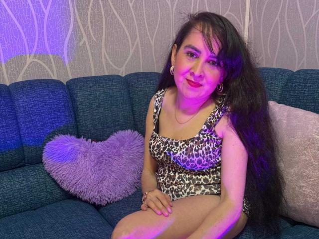 DianaLane with long black hair, wearing a leopard print dress, sitting on a blue couch with purple heart pillow, smiling with pink-purple light on her face.