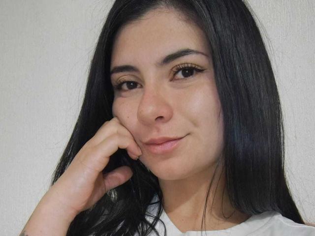 JeneKurz heeft lang zwart haar, bruine ogen met eyeliner, een kleine neus en een lichte glimlach. Ze leunt haar kin op haar hand en ziet er schattig uit..