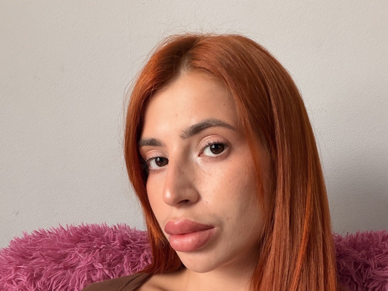 RosioRaschko has light brown skin, red straight hair, brown eyes, and puffy pink lips. She's looking slightly to the side with a neutral expression. Pink fuzzy pillow behind.