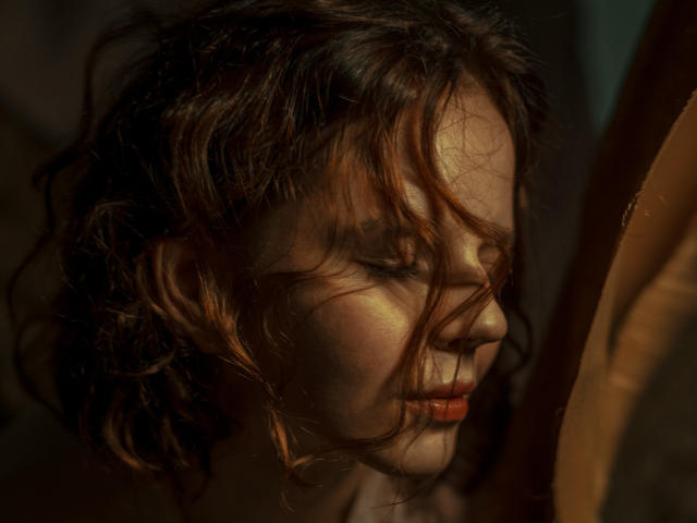Il viso di Adelheid, con i capelli castano ondulati e il rossetto rosso, è illuminato lateralmente, mostrando ombre delicate e un'espressione pensierosa, con gli occhi parzialmente chiusi..