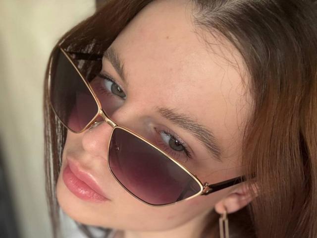 WhitneyLenort, avec des cheveux bruns, des yeux bleus et des lèvres roses, porte des lunettes de soleil sombres à monture dorée et regarde légèrement vers le haut avec une expression détendue. Ses fesses ne sont pas visibles..