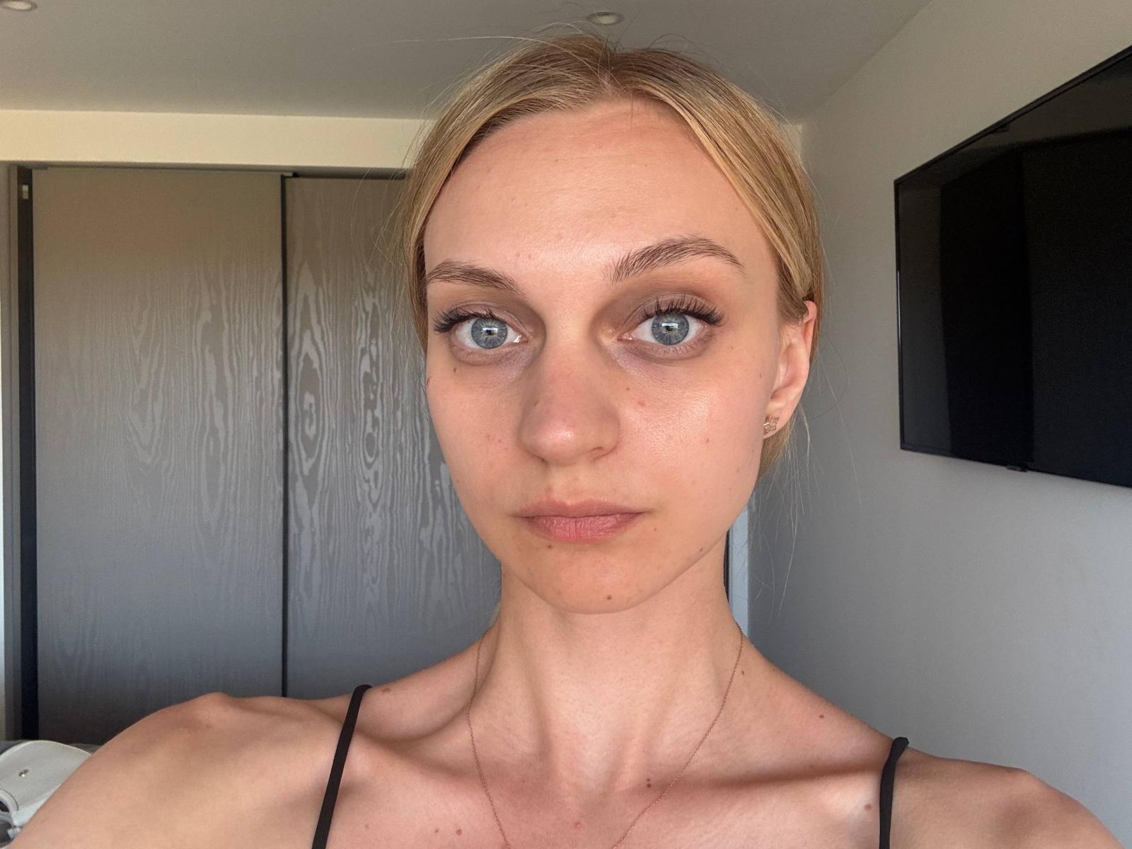 Earnestine Kendzierski tiene cabello rubio, ojos azules y una expresión seria, y lleva puesto un top negro con tirantes finos, que deja al descubierto sus hombros y lleva un collar pequeño..