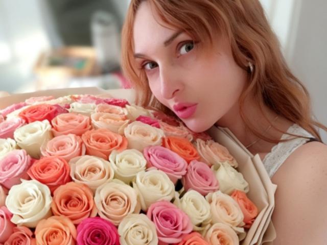 BelNell, avec des cheveux roux, tenant un bouquet de roses roses, oranges et blanches, regardant l'appareil photo avec du rouge à lèvres rose et une manche en dentelle blanche..