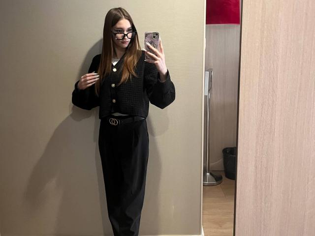 Candelaria Ortwein met lang bruin haar, een bril, een zwarte cardigan, een zwarte broek met hoge taille, die een telefoon vasthoudt, een spiegel-selfie maakt, en staat tegen een beige muur, terwijl ze naar de telefoon kijkt..