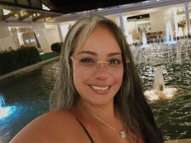 LailaCastillo with long, dirty blonde and black hair, wearing glasses and a black top, smiling at the camera, showing her tits, in a dimly lit, water-filled area.