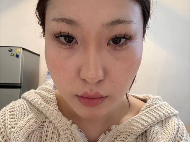 Le visage de MelinaBarne, qui regarde directement la caméra avec des lèvres pincées et de longs cils. Elle porte un sweat à capuche beige en tricot, et a une peau claire et des cheveux bruns foncés..