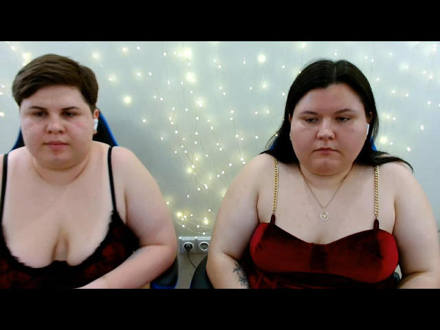 BeckyAndEllen, entrambe formose con grandi seni, indossano reggiseni di velluto neri e rossi, sono sedute, hanno capelli neri e castani, ci sono lucine decorative sullo sfondo, hanno espressioni neutre, c'è una cornice nera..