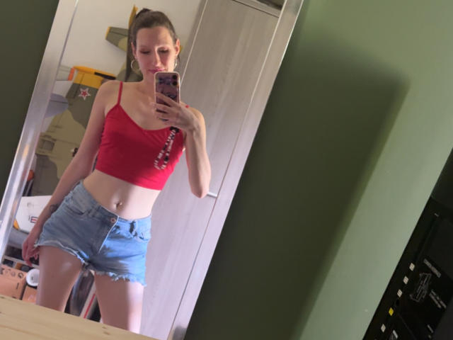 JadeFrenchKiss parada em frente a um espelho, vestindo uma blusa cropped vermelha e shorts jeans azuis, segurando um telefone, exibindo seu corpo esguio e tonificado e seus pequenos seios..