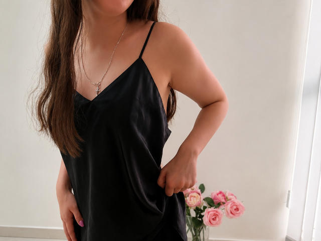 JoannaSia vêtue d'une nuisette en satin noir, montrant ses seins, avec un collier en argent, des cheveux bruns, des ongles roses, une main dans la poche, et des roses roses en arrière-plan..