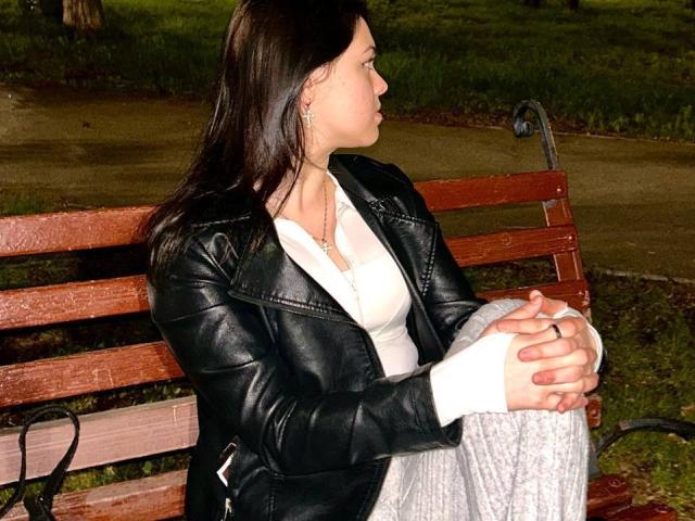 DelarySong sitzt auf einer hölzernen Bank, die Hände auf den Knien, und trägt eine schwarze Lederjacke, ein weißes Oberteil und eine graue Hose. Sie blickt zur Seite und hat dunkelbraunes Haar..