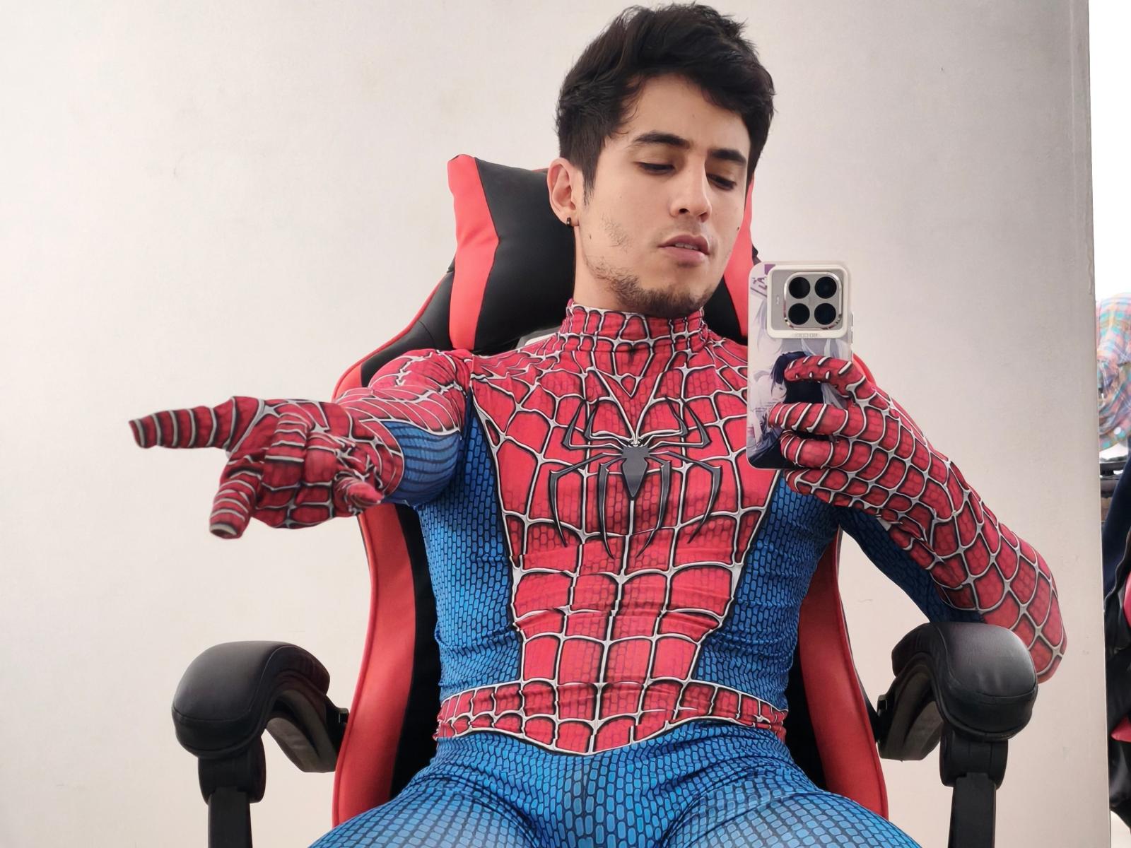 BrunoClaude in a Spider-Man suit, sitting on a red-black chair, pointing with his right hand, holding a smartphone in his left, looking down with a slight smirk.