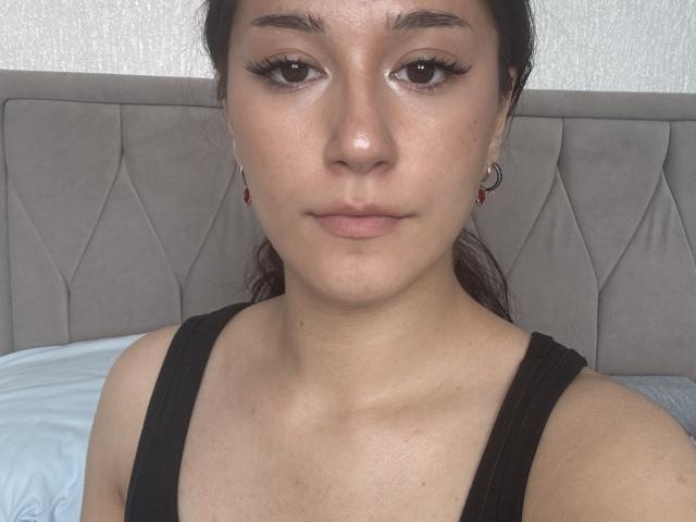 AngleZiak met een medium huidskleur, donker haar in een paardenstaart, bruine ogen, draagt een zwarte tanktop, subtiele make-up, rode oorbellen, kijkt recht in de camera met een neutrale uitdrukking..