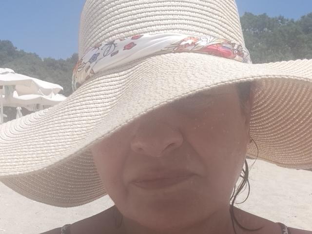 Esmeralda, com o rosto avermelhado pelo sol, usando um chapéu de palha bege grande com uma faixa floral e com o cabelo molhado, está em pé em uma praia de areia. Seu nariz é ligeiramente arrebitado..