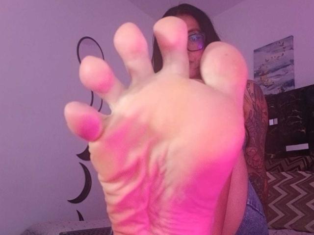 ZarayFeet's soles are in focus, pink-tinted toes and wrinkled skin, with tattooed arm and glasses, sitting in dim room. Her ass is partially visible.