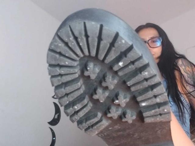 ZarayFeet holds a dirty, worn-out boot sole up to the camera, showing the gritty, black rubber tread with scattered dirt. She has long black hair and glasses.