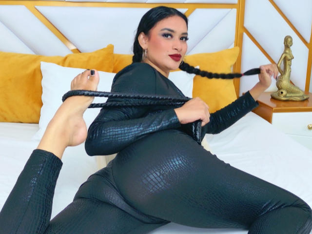 Adalina Vera mit schwarzen Zöpfen, trägt glänzende schwarze Hosen aus Schlangenleder, hält eine Peitsche, ihr großer Hintern ist im Fokus, auf einem Bett mit gelben Kissen und einer Buddha-Statue..