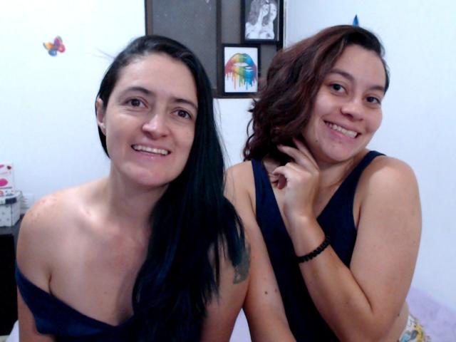 LunaVenus with black hair and small tits, and LunaVenus with brown hair, smiling together in a room. Both wear dark blue tank tops, showing some ass.