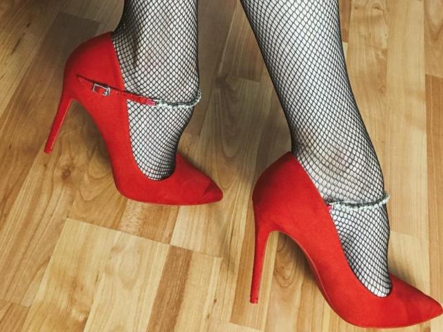 Nalini's red high heels with stiletto heels and fishnet stockings, standing on a wooden floor. Her feet are pointed, showing off the sexy, stylish outfit.