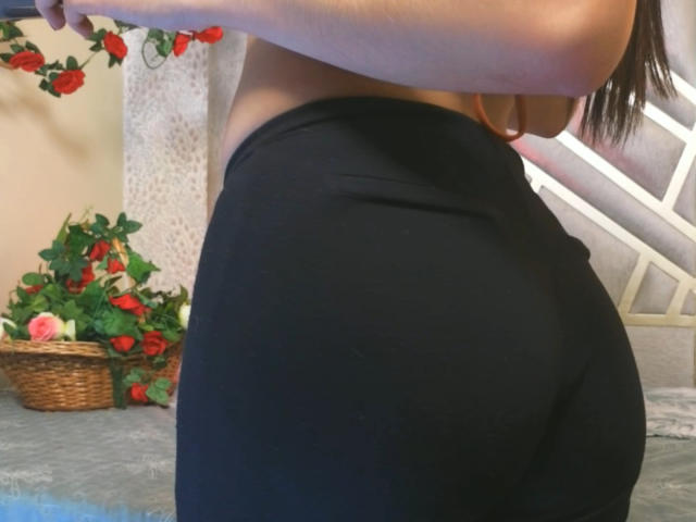 ChloeXk's butt in tight black shorts, showing a curvy, firm ass. She's wearing a top, and her long brown hair is visible. Red roses in the background.